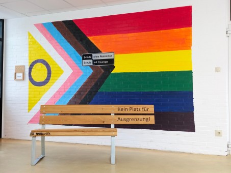 Bank mit Text "Kein Platz für Ausgrenzung" vor dem Wandbild "Schule gegen Rassismus - Schule mit Courage". Ein Teil der Sitzbank fehlt, damit ein Rollstuhl dort stehen kann..