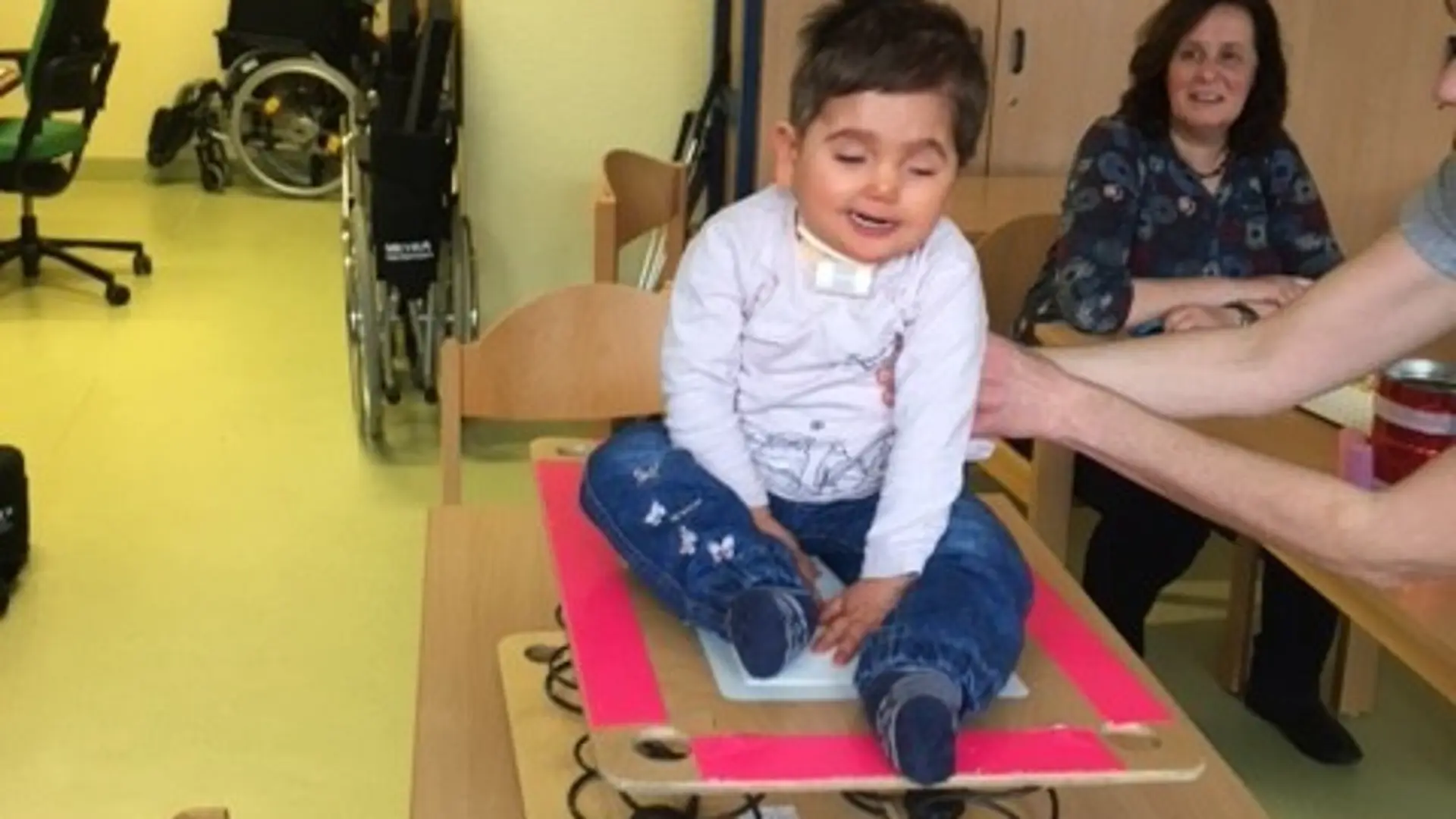 Ein Kleinkind sitz auf einem gefederten Brett und versucht, das Gleichgewicht zu halten. Eine Frau stützt es mit den Händen.