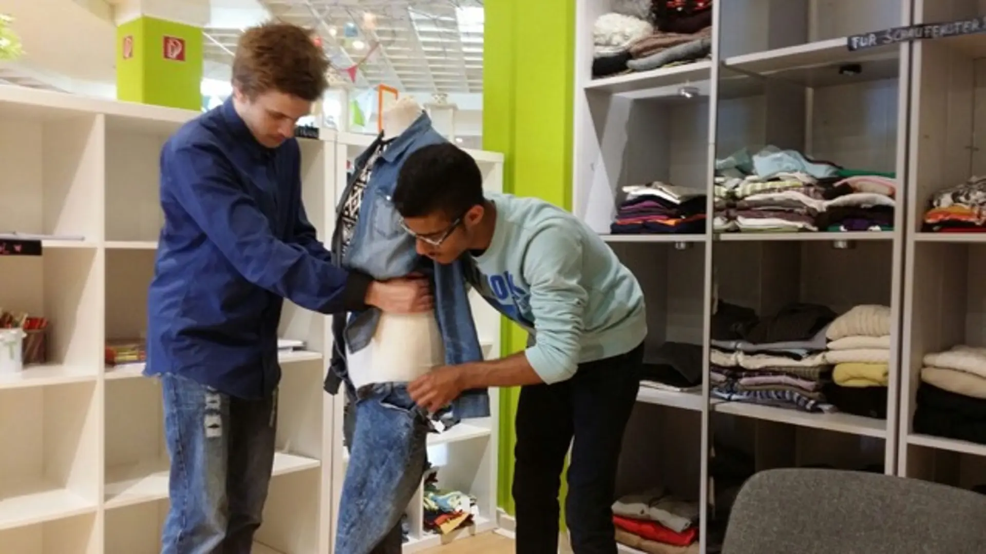 Zwei Jugendliche ziehen einer Schaufensterpuppe Jeanskleidung an. Im Hintergrund ein Raum mit Regalen, in denen Kleidung gestapelt ist.