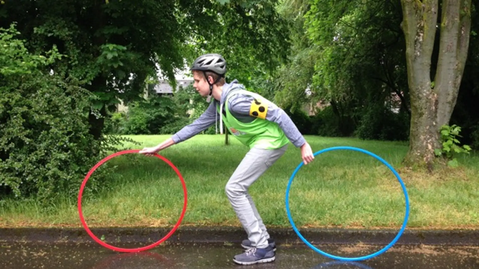 Man sieht einen Jungen von der Seite. Er steht gebeugt und hält einen Turnreifen vor sich, einen zweiten hinter sich. Das sieht so aus, als würde er auf einem Fahrrad sitzen..