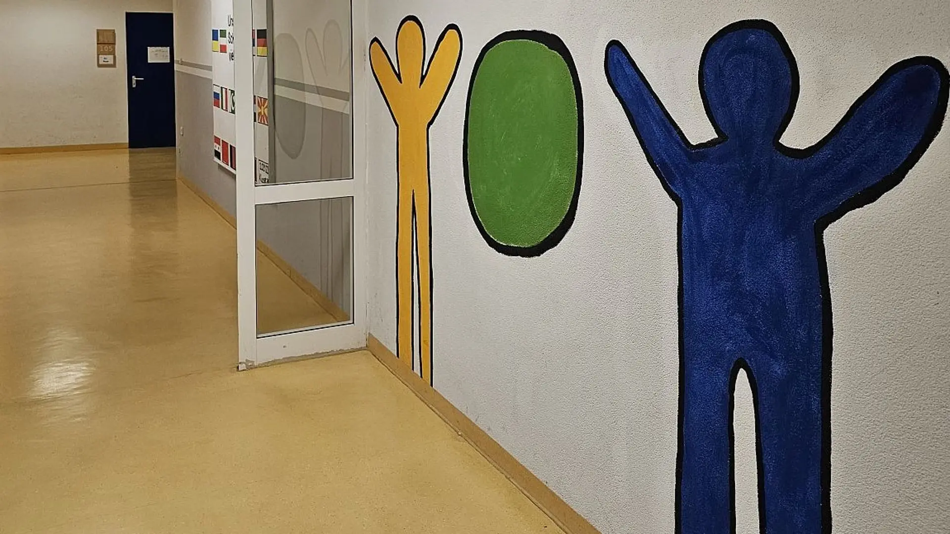 Ein langer Flur. An der Wand zwei gemalte Figuren im Stil von Keith Haring. Sie strecken die Arme nach oben: Zwischen ihnen ist ein Ball gemalt.