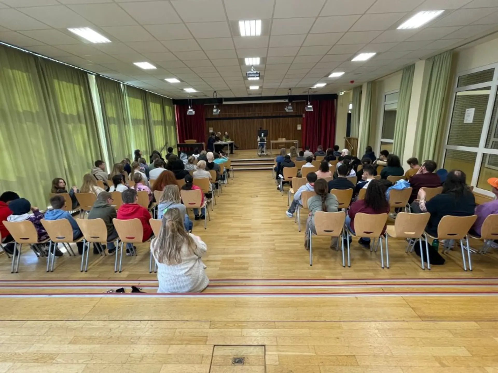 Eine Gruppe Menschen von hinten: Sie sitzen auf Stühlen in einem großen Raum und schauen nach vorne, wo auf einer Bühne Menschen etwas vorlesen.