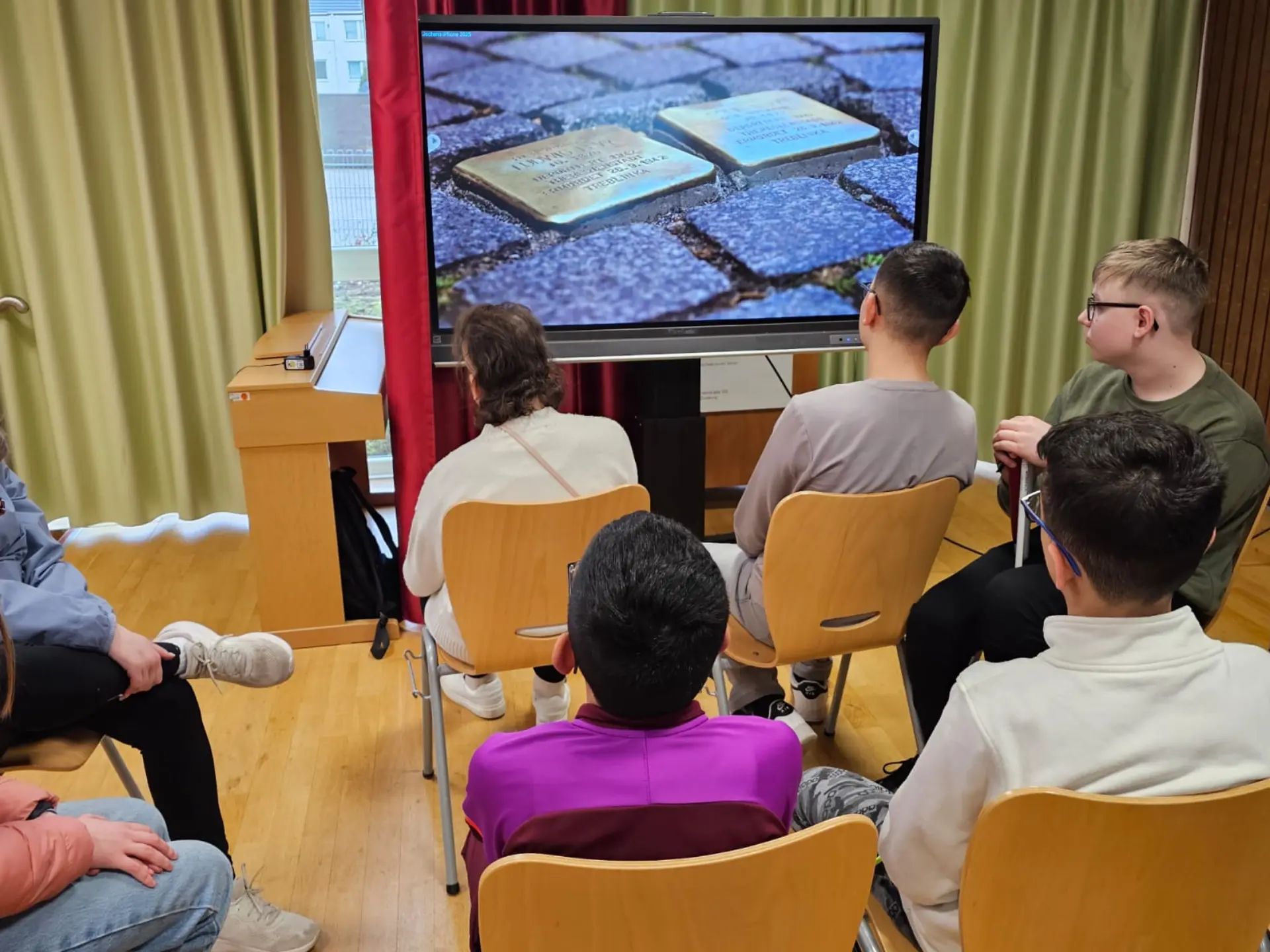 Schüler:innen sitzen auf Stühlen vor einem Smartboard und schauen einen Film zum Thema Stolpersteine  