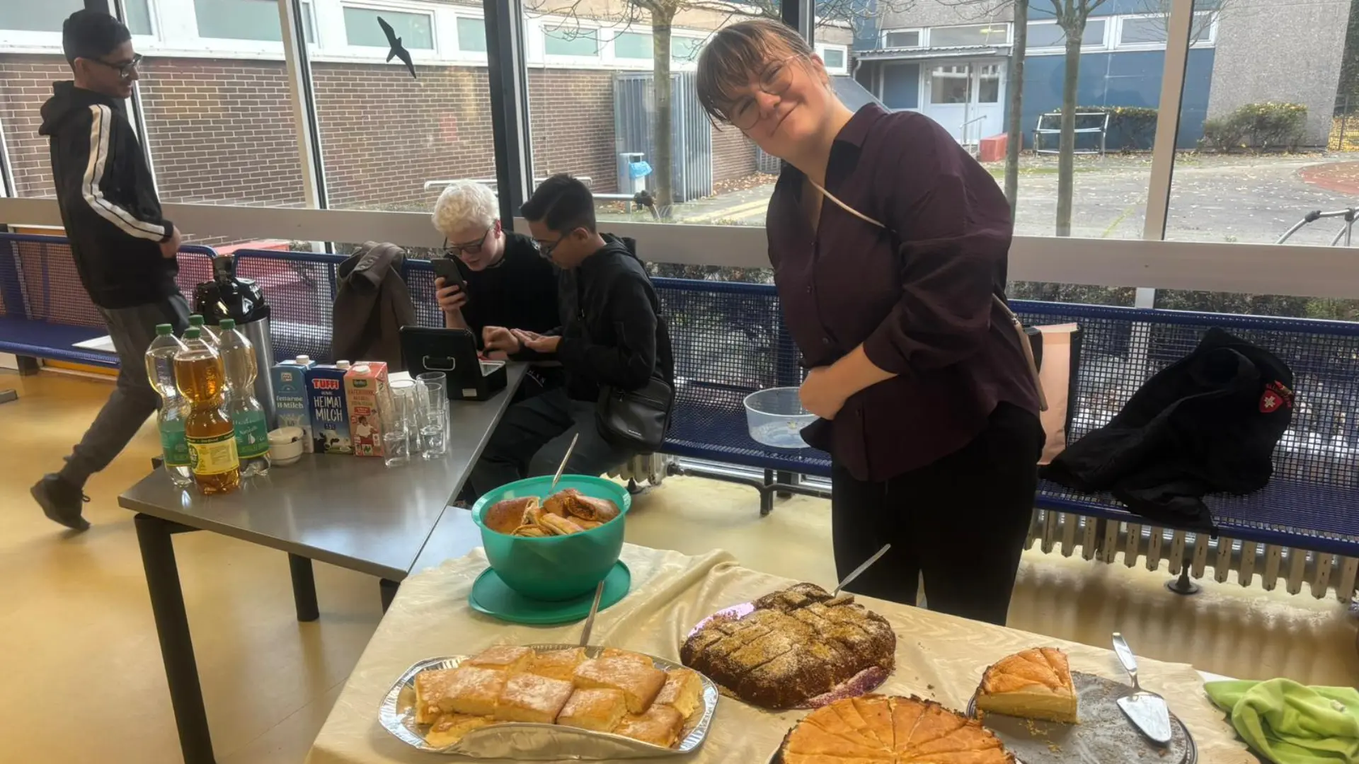 Eine Frau steht hinter einem Tisch, auf dem verschiedene Kuchen stehen. Sie lächelt freundlich. Auf einem weiteren Tisch stehen Getränke. Dahinter sind drei Jugendliche, die die Getränkeausgabe betreuen.