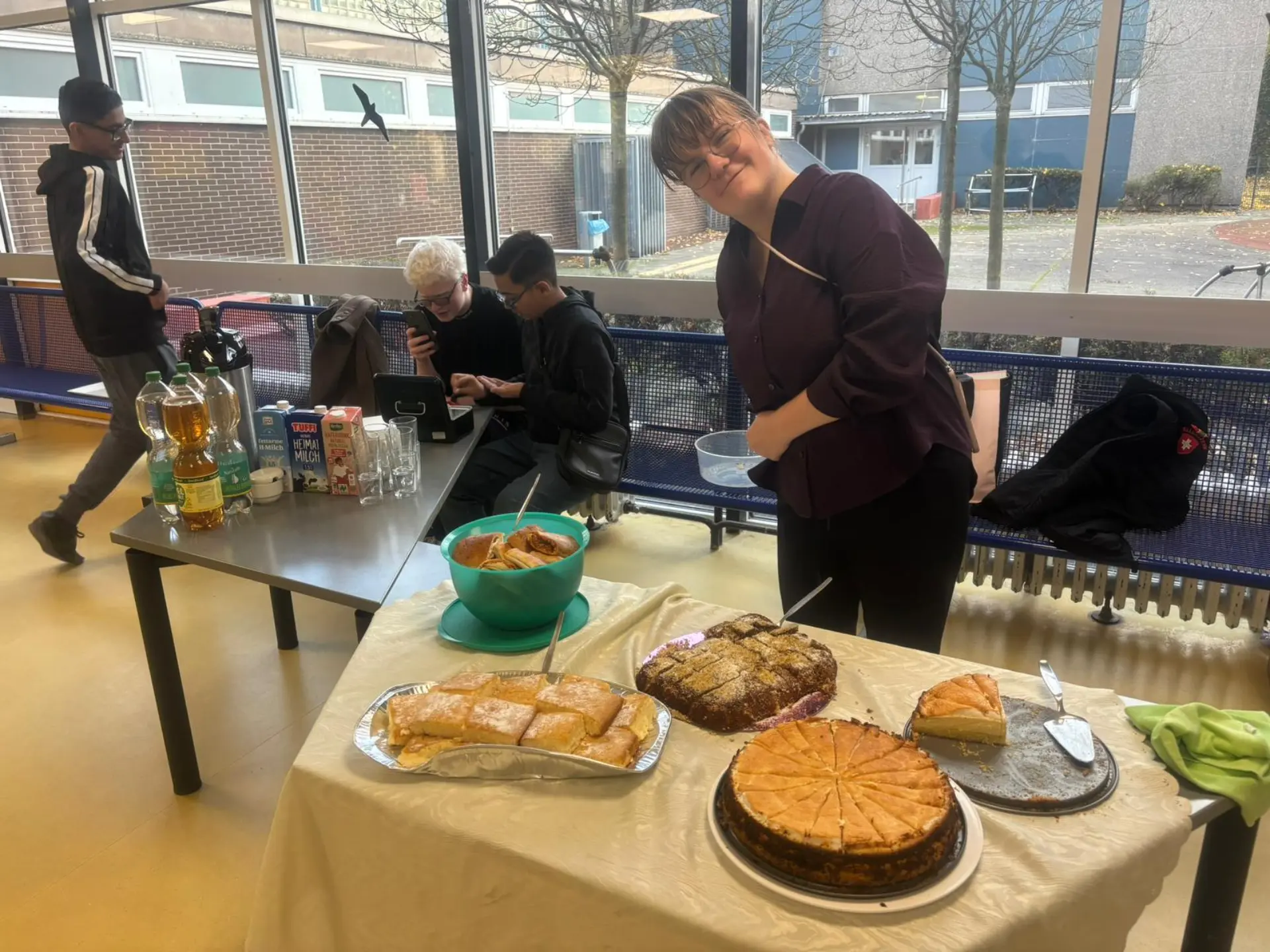 Eine Frau steht hinter einem Tisch, auf dem verschiedene Kuchen stehen. Sie lächelt freundlich. Auf einem weiteren Tisch stehen Getränke. Dahinter sind drei Jugendliche, die die Getränkeausgabe betreuen.