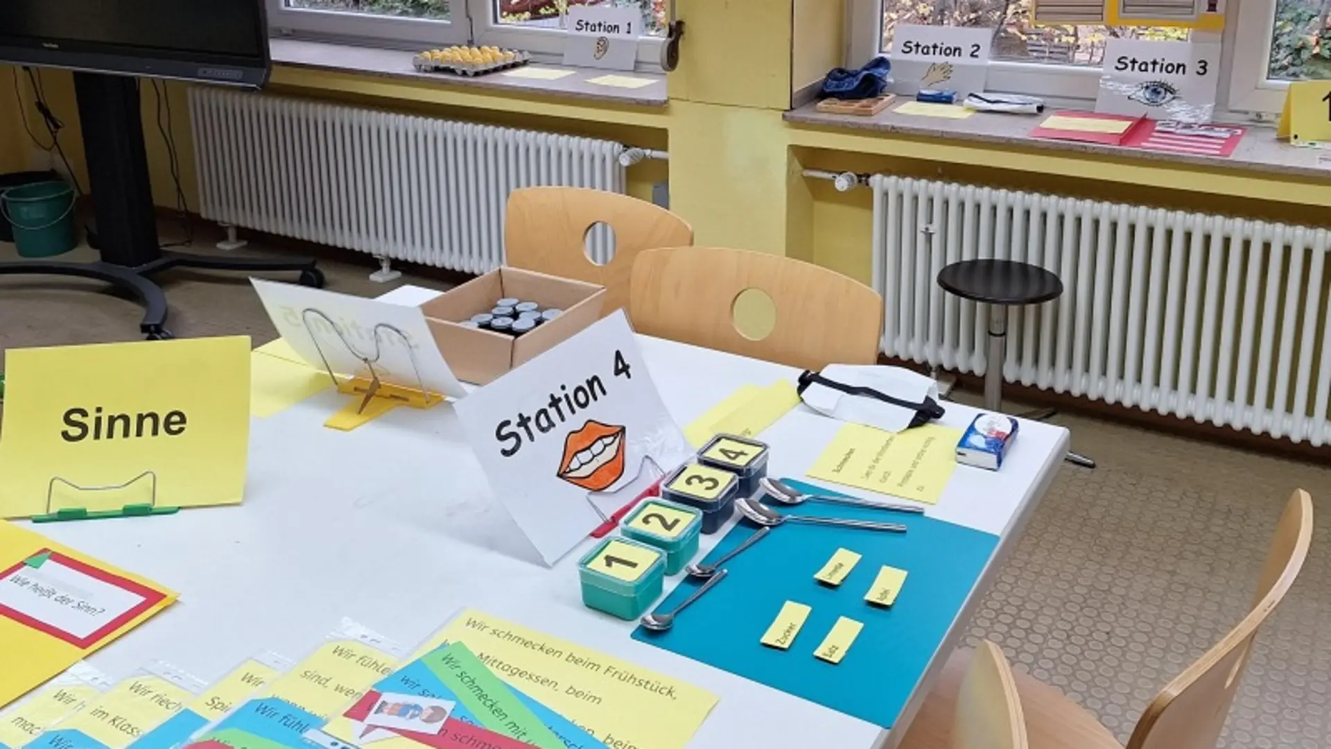 In einem Klassenraum steht ein Tisch mit Material zum Thema "Sinne", zum Beispiel kleine Dosen mit  den Zahlen 1-4 beschriftet und Löffel. An den Fenstern hängen Plakate zum Thema "Sinne". Auf dem Fensterbrett liegen weitere Materialien zu den Stationen 1, 2 und 3.