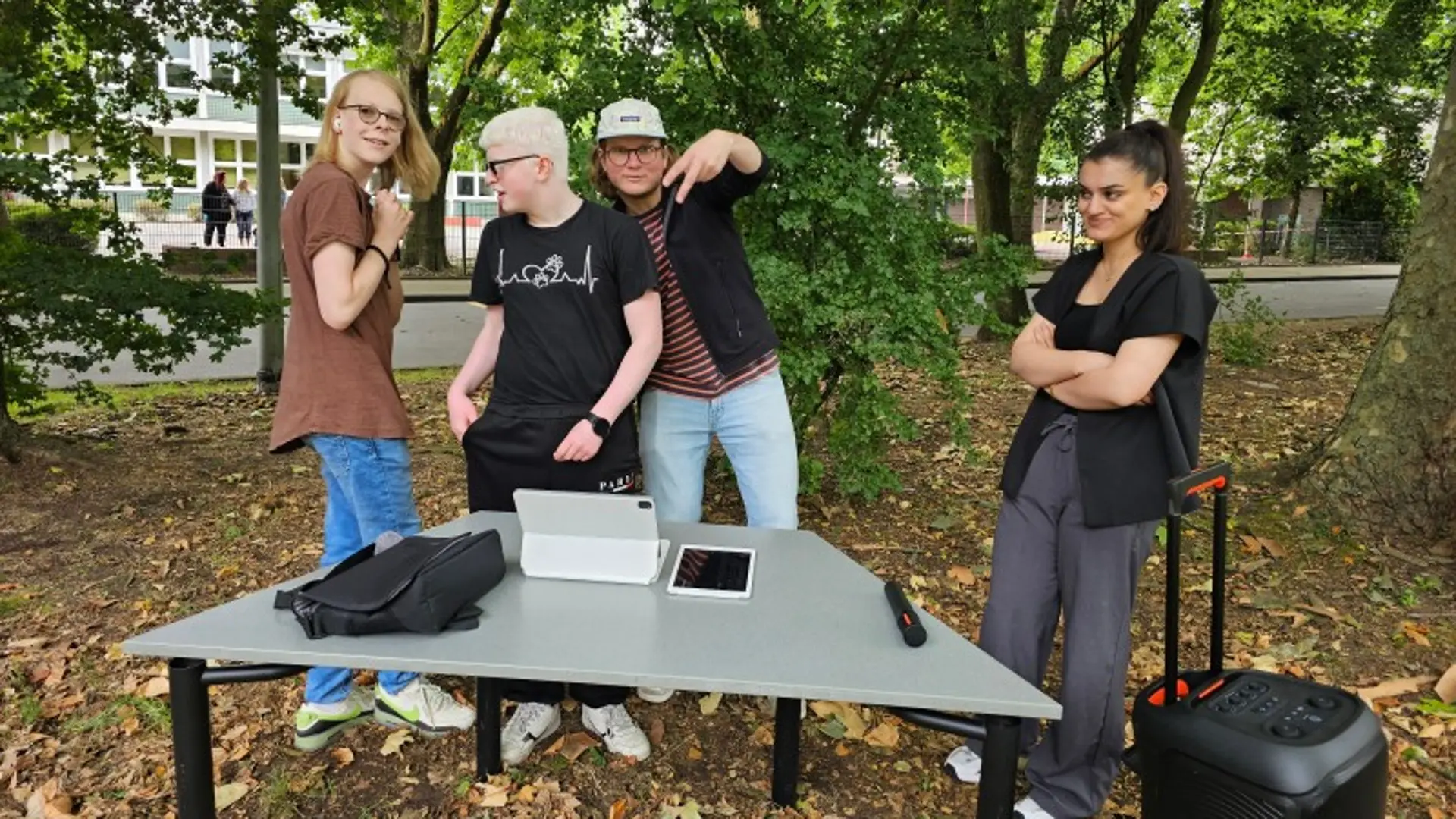 Vier Jugendliche tanzen unter Bäumen. Vor ihnen steht ihnen ein Tisch mit einem Notebook, daneben ein mobiler großer Lautsprecher.