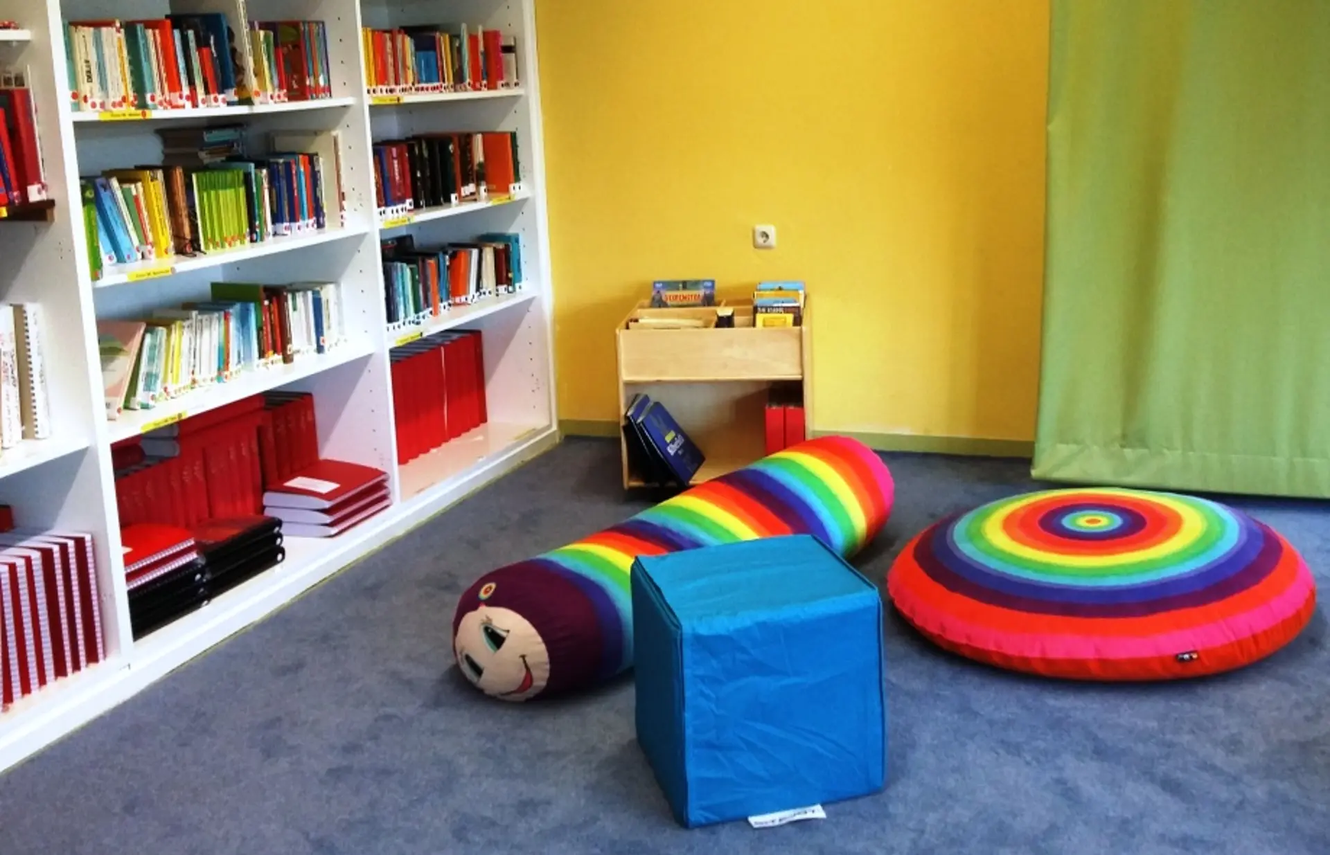 Ein freundlicher Raum mit Bücherregalen, blauem Teppich und großen, regenbogenfarbenen Kissen auf dem Boden