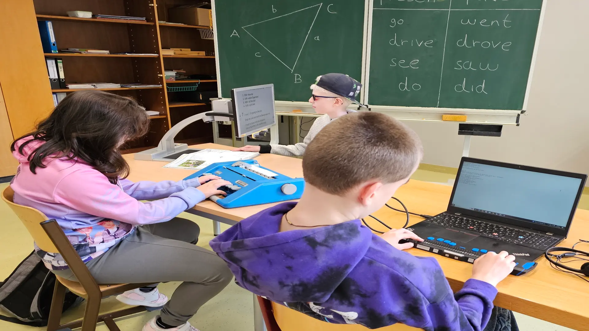 Zwei Schüler und eine Schülerin arbeiten im Klassenzimmer am Computer, Brailledrucker und mit Vergrößerungslupe. Im Hintergrund eine grüne Tafel.