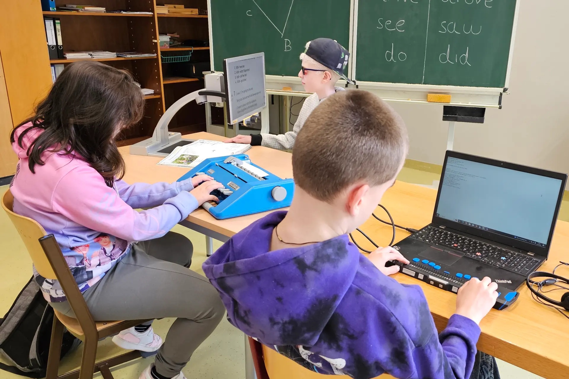 Zwei Schüler und eine Schülerin arbeiten im Klassenzimmer am Computer, Brailledrucker und mit Vergrößerungsdings. Im Hintergrund eine grüne Tafel.
